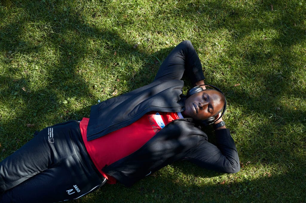 Assan Jallow liegt in Sportbekleidung, mit geschlossenen Augen und mit Kopfhörern auf dem Rasen