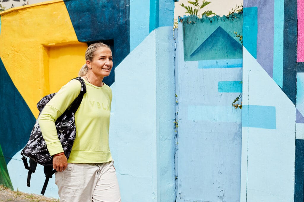 Nikki Adler trägt ein hellgelbes Shirt und einer schwarzen Tasche vor einer hellblauen Wand.