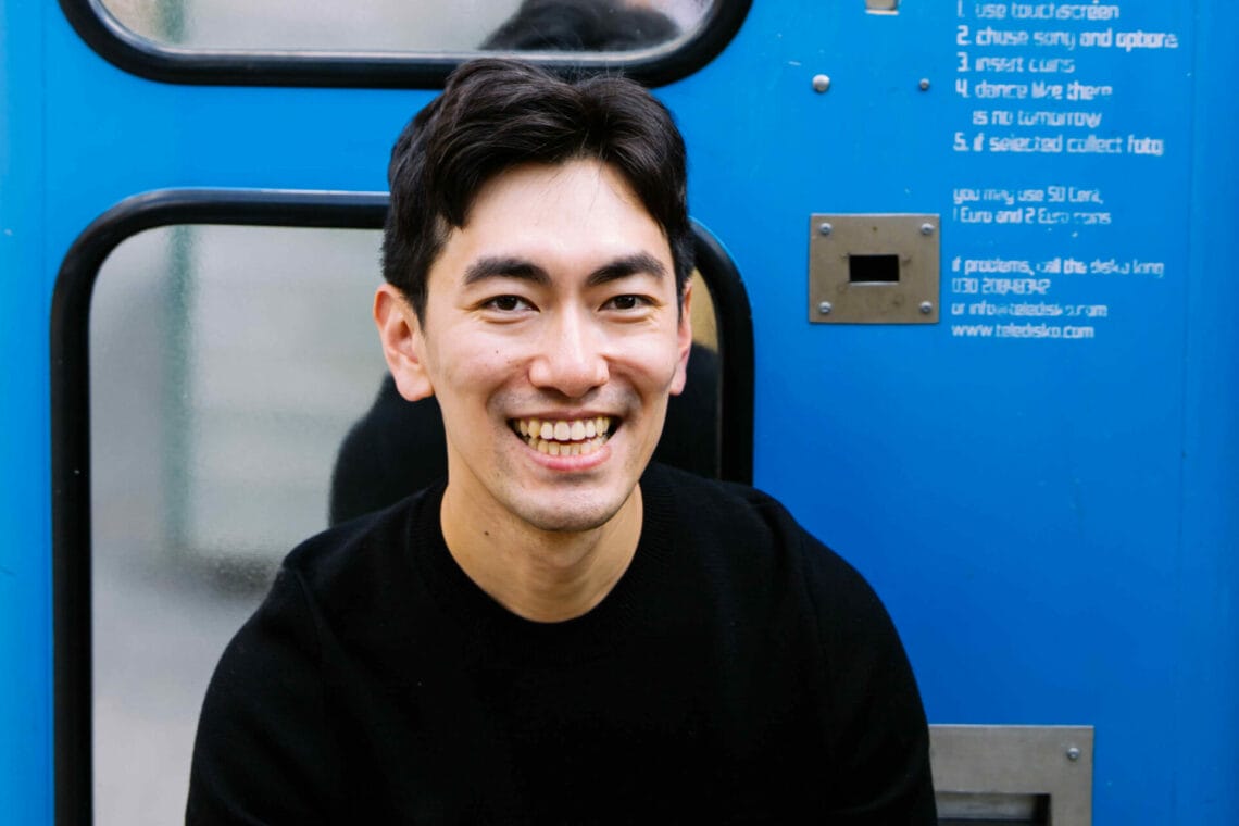 Sung UN Gang sitzt im schwarzen Sweatshirt vor einem blauen Fotoautomaten