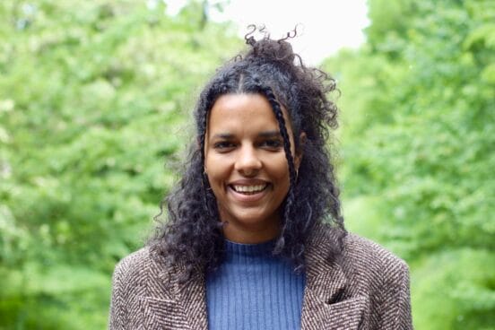 Autorin Alice Hasters trägt einen blauen Wollpulli unter einem beigen Sakko und lächelt in die Kamera. Im Hintergrund ist das Grün von Bäumen zu sehen.