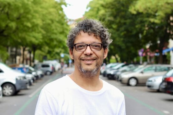 Atif Hussein blickt im weißen T-Shirt lächelnd in die Kamera. Er steht auf der Straße, im Hintergrund sind links und rechts Autos zu sehen