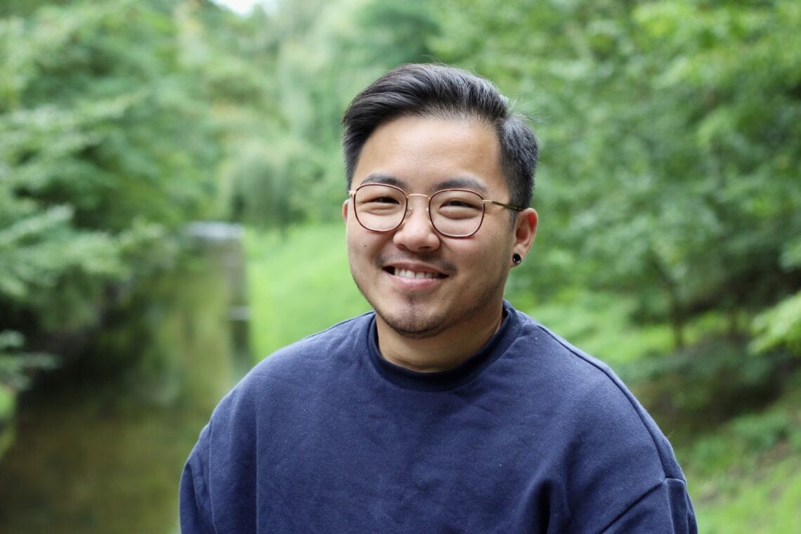 Duke Duong steht im blauen Sweatshirt vor grünem Bäumen