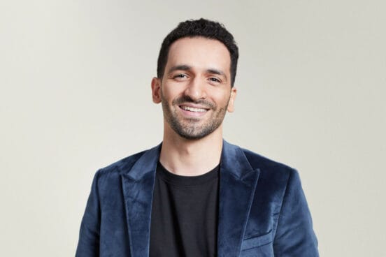 Schauspieler Hssan Akkouch steht lächelnd im schwarzen Shirt und blauem Sakko vor einer beigen Wand.