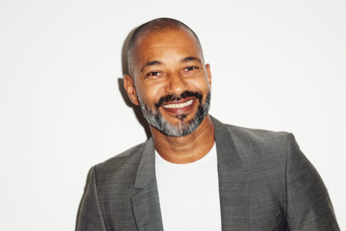 Schauspieler Tyron Ricketts lächelt im weißen Shirt und grauem Sakko in die Kamera