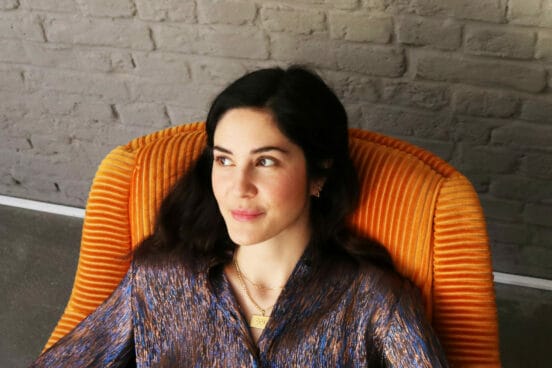 Yelda Türkmen trägt eine dunkle Bluse und sitzt in einem orangefarbenenen Sessel. Sie guckt zur Seite und lächelt.