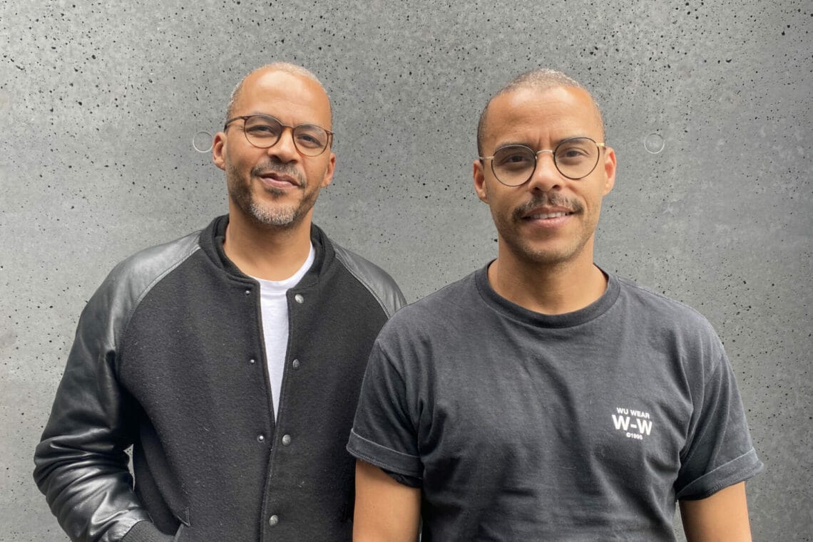 Jeffrey Lisk trägt ein weißes Shirt und darüber eine Schwarz-graue kragenlose Collegejacke (links) und Dennis Lisk aka Denyo ein schwarzes T-Shirt. Beide setehn nebeneinander und sind halbhoch zu sehen.