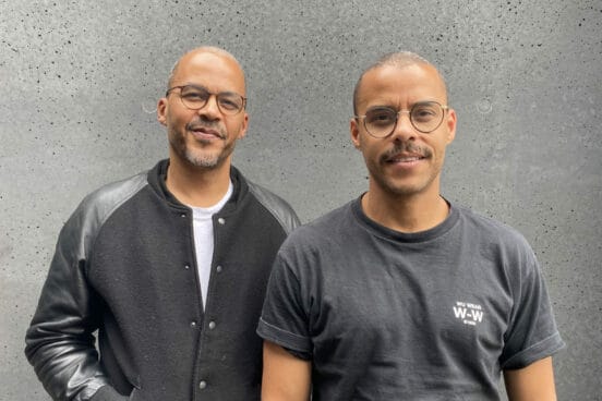 Jeffrey Lisk trägt ein weißes Shirt und darüber eine Schwarz-graue kragenlose Collegejacke (links) und Dennis Lisk aka Denyo ein schwarzes T-Shirt. Beide setehn nebeneinander und sind halbhoch zu sehen.