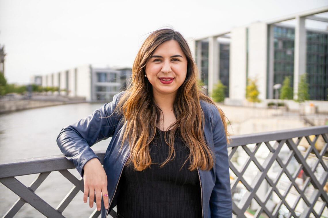 MIna Saidze steht auf einer Brücke im Berliner Regierungsviertel. HInter ihr ist Wasser zu sehen. Sie trägt ein schwarzes Top und darpber eine schwarze Lederjacke.
