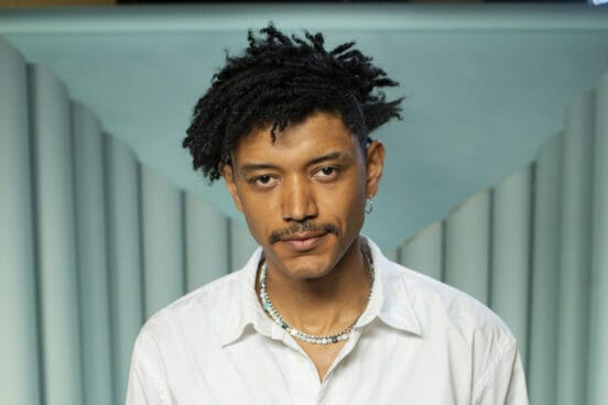 Don Pablo Mulemba trägt ein weißes Hemd und zwei Ketten, lächelt in die Kamera und steht vor einem grünen Hintergrund in Studio-Atmosphäre.