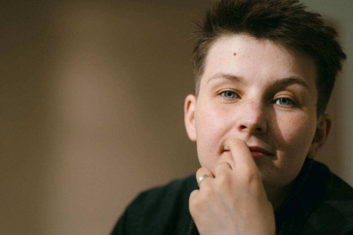 Porträtbild von Queermed-Gründer*in Samson Grzybek schaut in die Kamera, der linke Zeigefinger kratzt am Mund