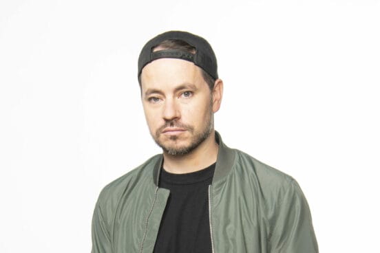 Raffi Balboa im schwarzen Shirt und olivgrüner Jacke ohne Kragen und schwarzen Käppi rückwärts schaut ernst in die Kamera vor weißem Hintergrund