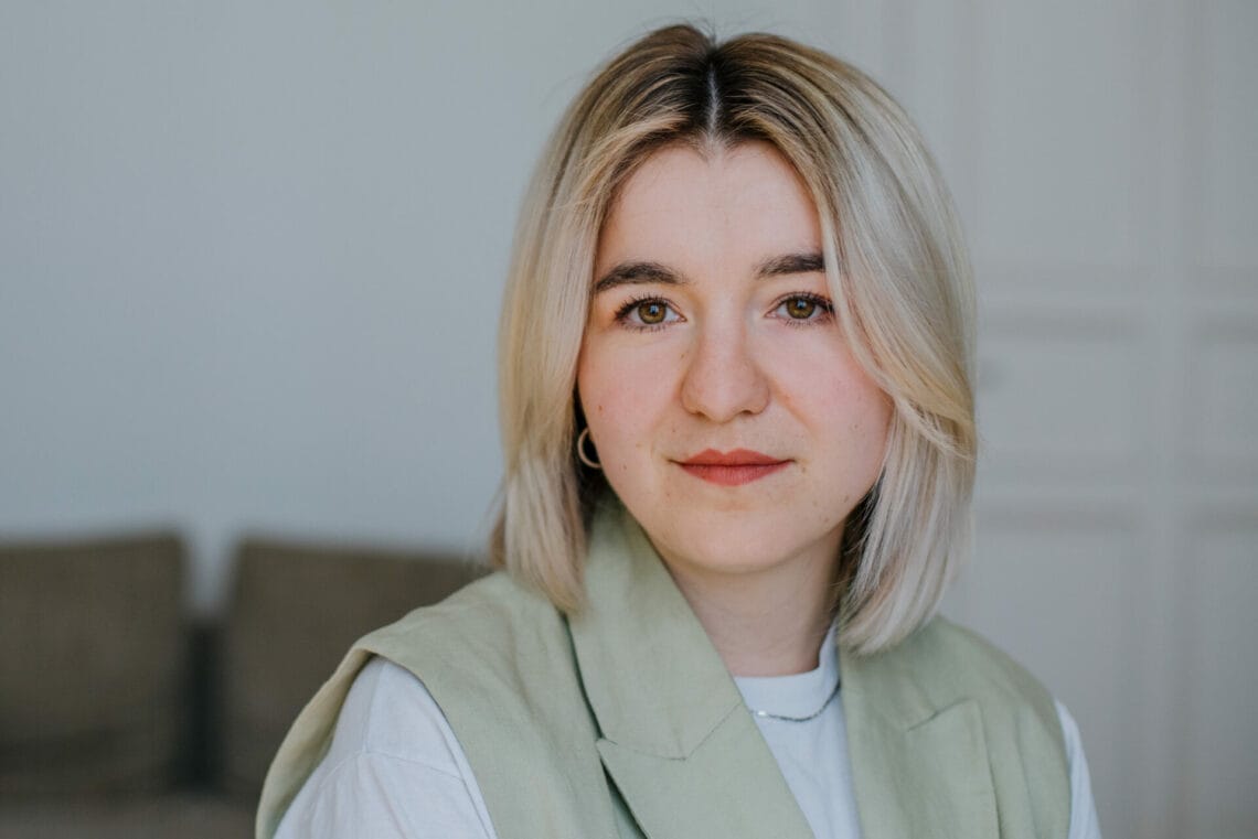 Maria Popov trägt ein weißes Shirt und eine mintgrüne Weste. Sie schaut in die Kamera