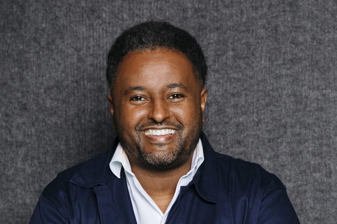 Fetsum trägt ein weißes Hemd und eine blaue Jacke darüber vor einem grauen Hintergrund und lächelt in die Kamera.