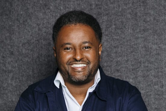 Fetsum trägt ein weißes Hemd und eine blaue Jacke darüber vor einem grauen Hintergrund und lächelt in die Kamera.