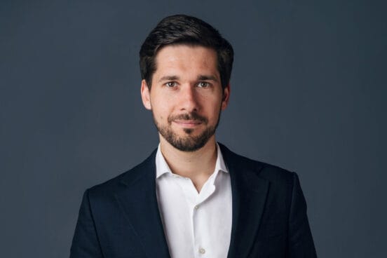 Vassili Golod steht im weißen Hemd und schwarzen Sakko vor einer dunkeln Wand und schaut frontal in die Kamera