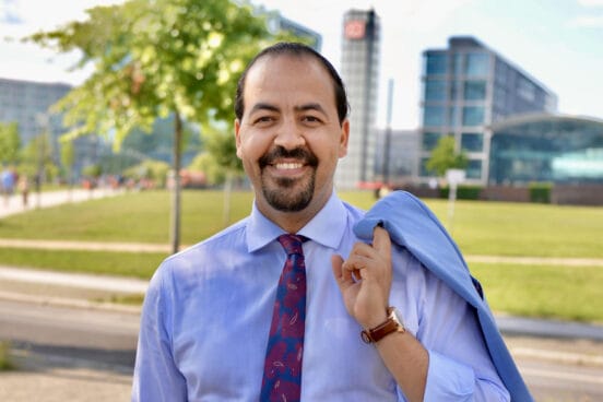 Mohammed El Ouahhabi trägt ein hellblaues Hemd, eine rotblaue Krawartte und ein blaues Sakko über der chulter. Die Sonne scheint und im Hintergrund ist der Berliner Hauptbahnhof zu sehen