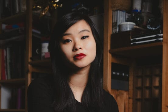 Nhi Le trägt ein schwarzes Oberteil und schaut ernst in die Kamera. Hinter ihr steht ein Regal.