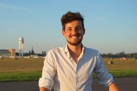 Ingwar Perowanowitsch trägt ein hochgekrempeltes, weißes Hemd und lächelt in die Kamera. Er steht aucf einer freien Fläche mit Rasen im Hintergrund in der Abendsonne.