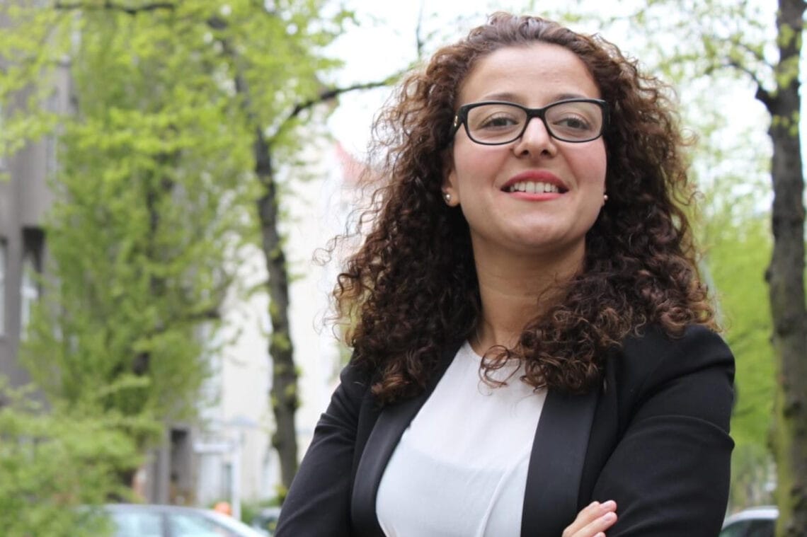 Jouanna Hassoun steht draußen auf der Straße. Sie trägt ein weißes Shirt und einen schwarzen Blazer und lächelt in die Kamera.