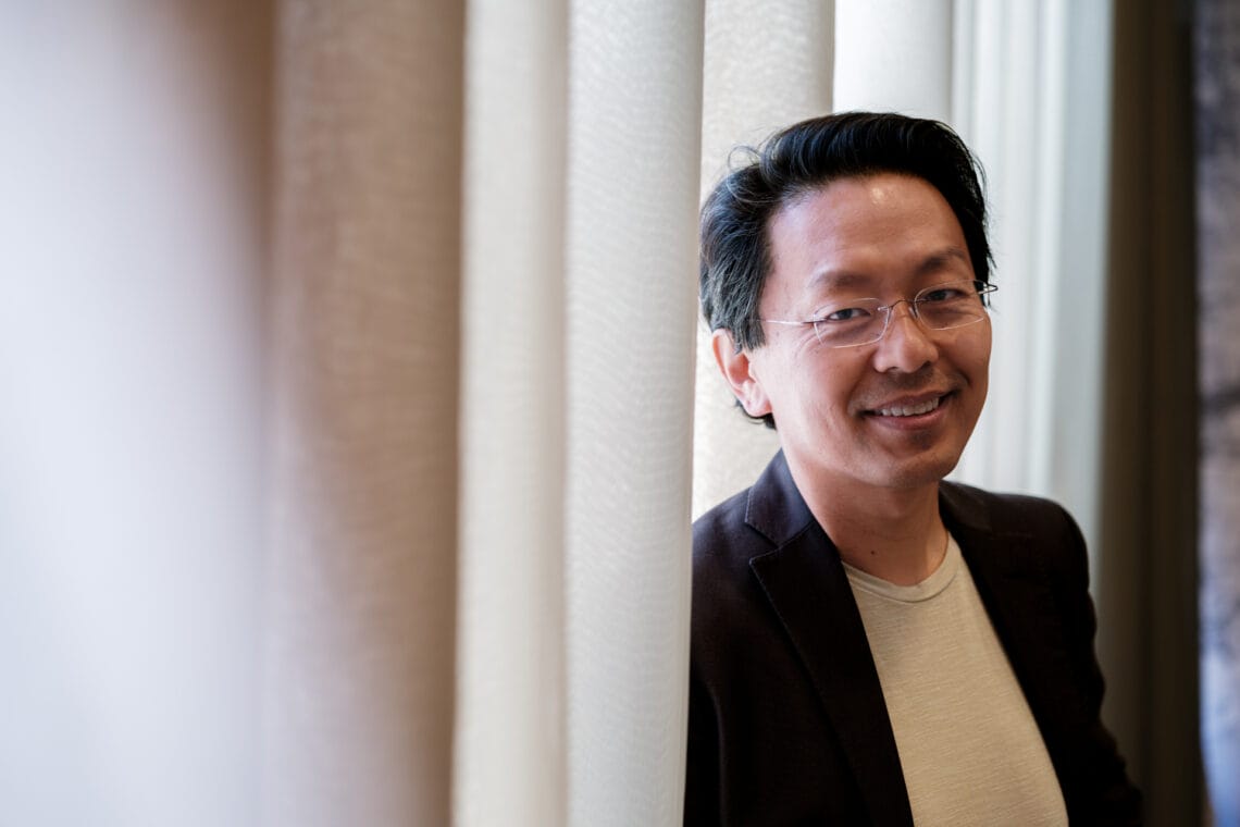 Chan-Jo Jun trägt ein dunkles Jackett über einem hellen Shirt. Er steht lächelnd vor einem Fenster mit Bürovorhang.