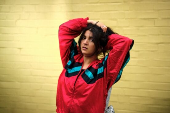 Roxana Samadi trägt eine rote Trainingsjacke mit Streifen, hält ihre Arme über den Kopf, als wolle sie ihre Haare binden. Sie steht vor einer gelben Wand.