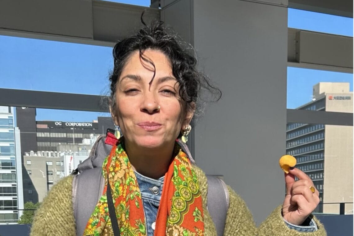 Yalda Ghods trägt eine olivegrüne Wolljacke, einen Rucksack und ein buntes Tuch um den Hals. Sie lächelt mit geschlossenem Mund in die Sonne und hält einen Snack in der Hand.