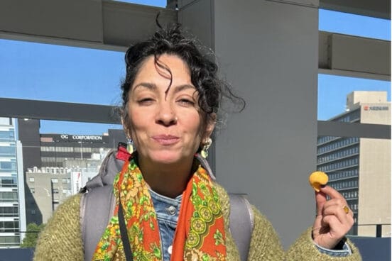 Yalda Ghods trägt eine olivegrüne Wolljacke, einen Rucksack und ein buntes Tuch um den Hals. Sie lächelt mit geschlossenem Mund in die Sonne und hält einen Snack in der Hand.