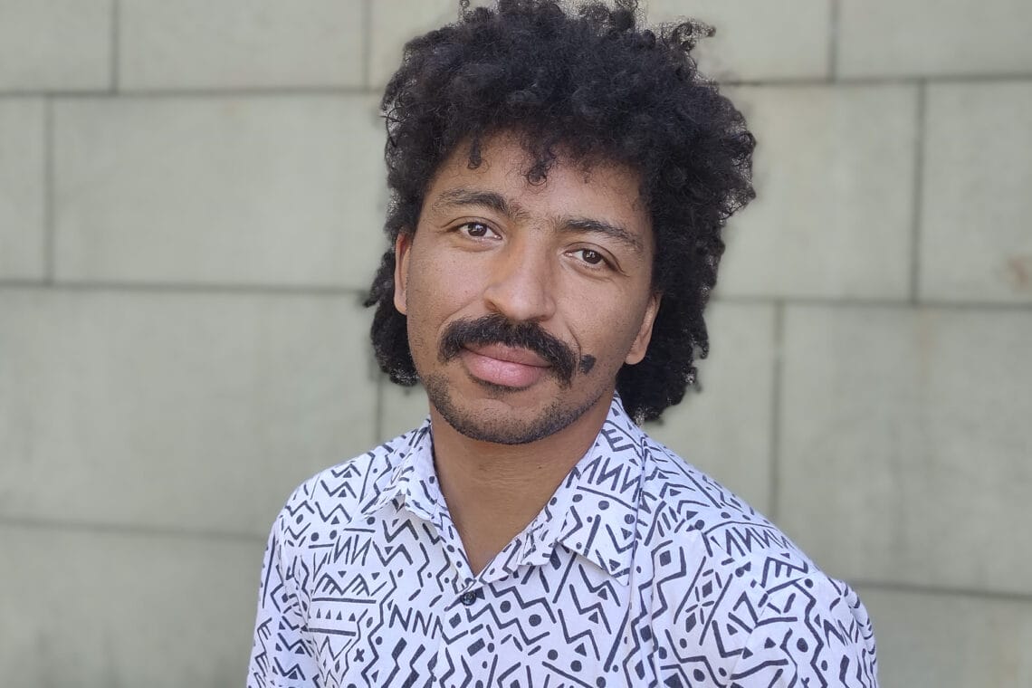 David Zabel steht vor einer beige-grauen Steinwand und trägt ein weißes Hemd mit schwarzem Muster.