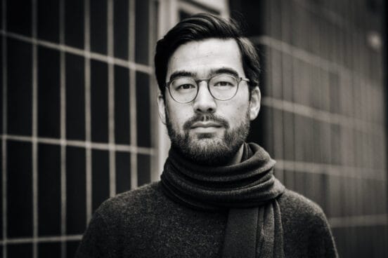 Yannic Han Biao Federer trägt einen dunklen Wollpullover und einen Schal. Er schaut ernst in die Kamera, hinter ihm ist ein Gitter zu sehen. Das Bild ist in Schwarz-Weiß.