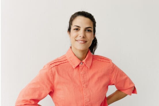 Clarissa Corrêa da Silva trägt ein pastell orange farbenes Hemd, die Hände in die Hüfte gestemmt und lächelt in die Kamera. Die Wand hinter ihr ist weiß.
