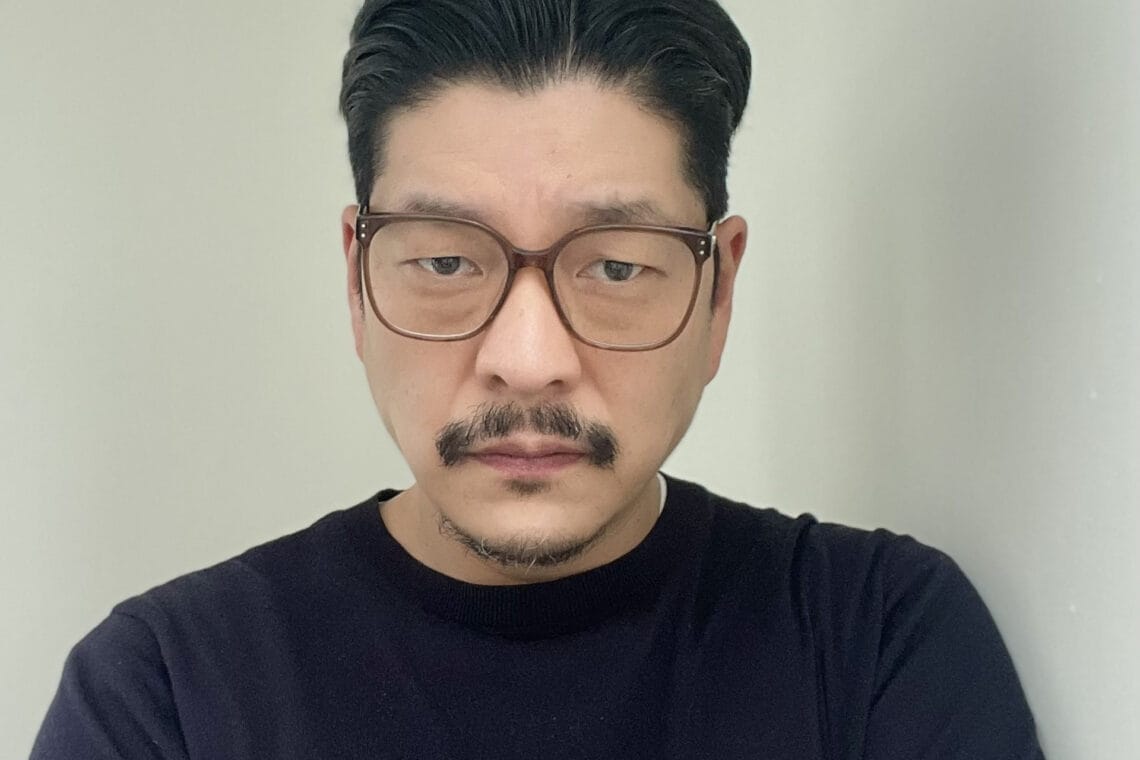 Bill Yom trägt einen schwarzen Sweater vor hellem Hintergrund und schaut ernst in die Kamera.