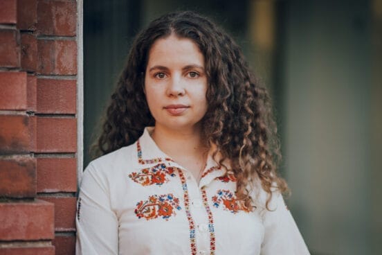 Militsa Tekelieva trägt eine weiße Bluse mit roten Ornamenten und lehnt an einer roten Steinwand.