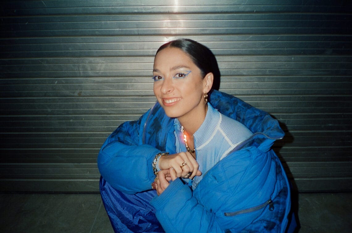 Tamara Güçlü aka Tam hockt im Blitzlicht mit himmelblauer Jacke vor einem Garagentor und lächelt.